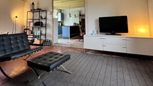 Een moderne woonkamer met een zwarte leren bank, een bijpassend poefje, een flatscreen televisie op een wit meubelstuk en een boekenkast vol boeken en decoratieve objecten.