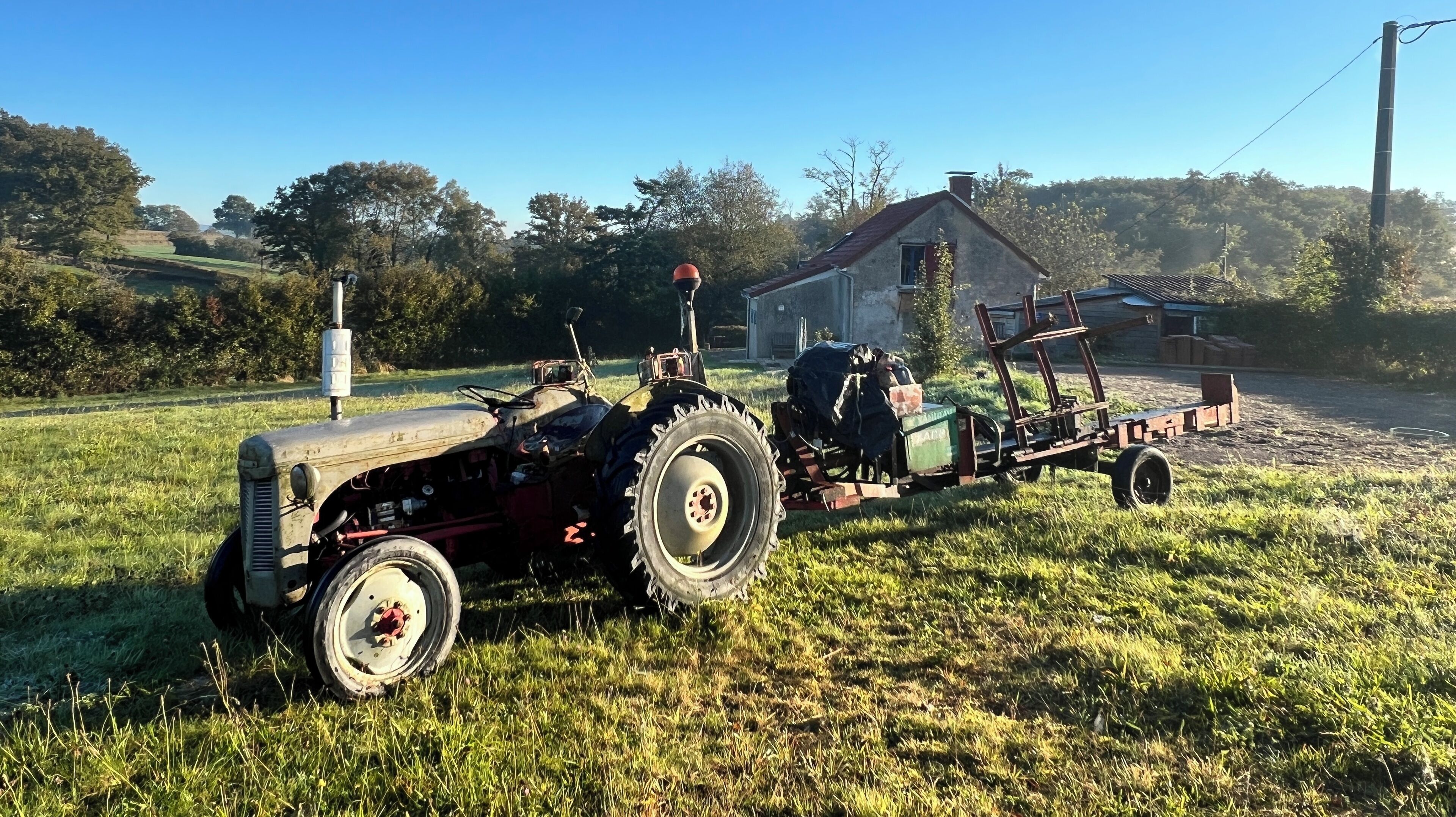 A vintage tractor with a trailer attached, parked in a grassy field with a house in the background.