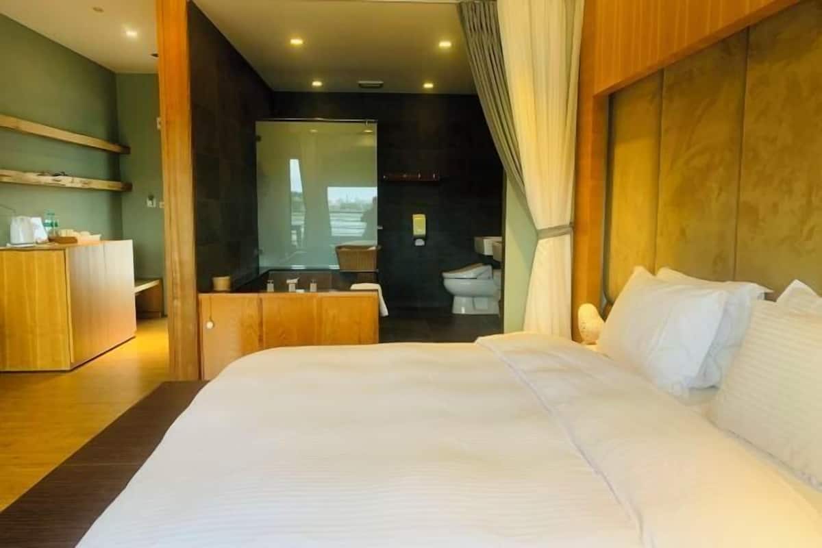 A modern hotel room with a large bed, a wooden headboard, a bathroom with a toilet and sink, and a view through a glass door.