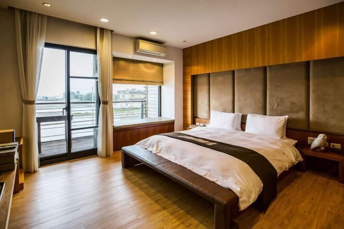 A hotel room with a large bed, a bench, a nightstand, a window with curtains, and a view of the outside.