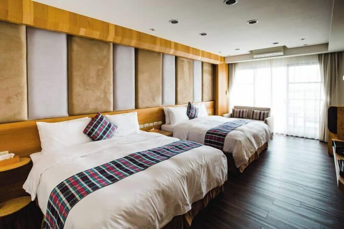 A hotel room with two beds, a wooden headboard, a large window with curtains, and a flat-screen TV on a wooden stand.