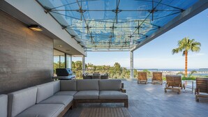A modern outdoor seating area with a glass ceiling, a barbecue grill, and a view of the coastline.