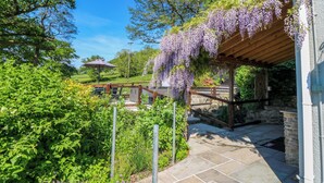 Een tuin met een pergola vol paarse bloemen, een kiezelpad en weelderige begroeiing.