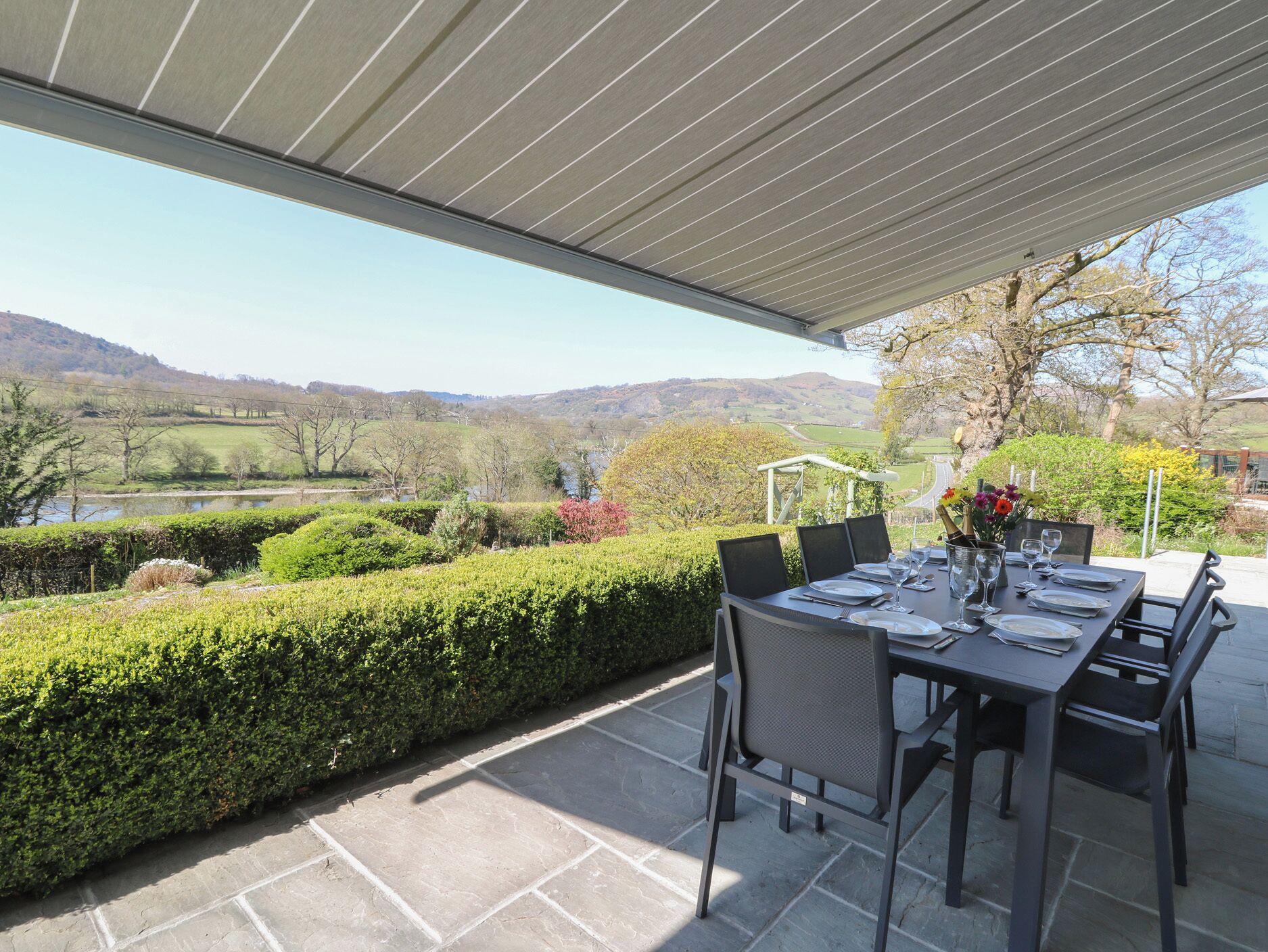 An outdoor dining area with a table set for four, overlooking a scenic landscape with hills and a body of water.