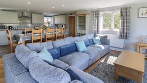 Een moderne keuken met witte kasten, een eettafel voor vier personen en een grote grijze hoekbank met blauwe kussens.