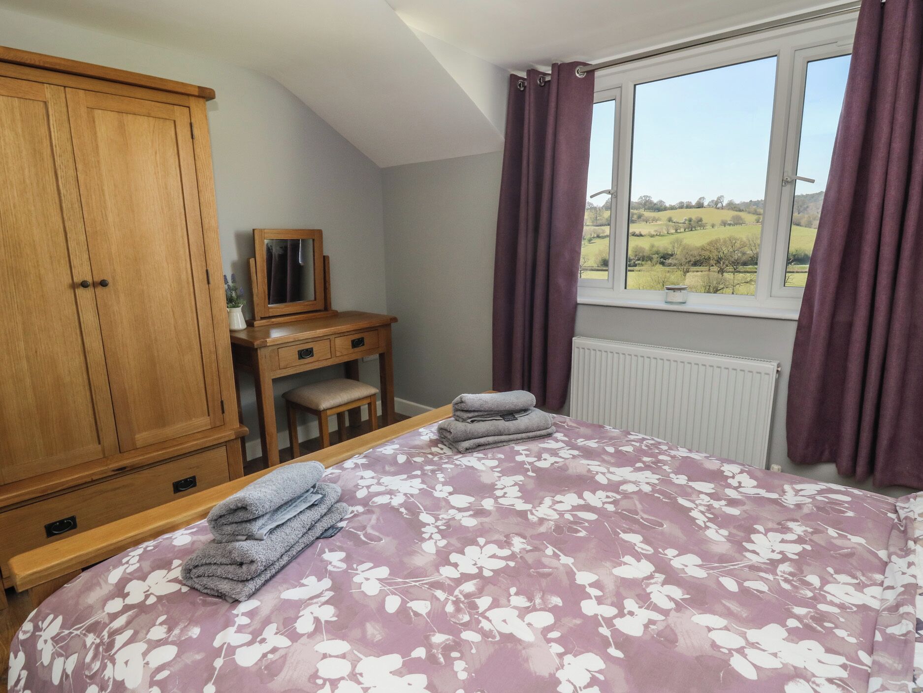 A bedroom with a bed, wooden wardrobe, a desk with a mirror, and a window with a view of the countryside.