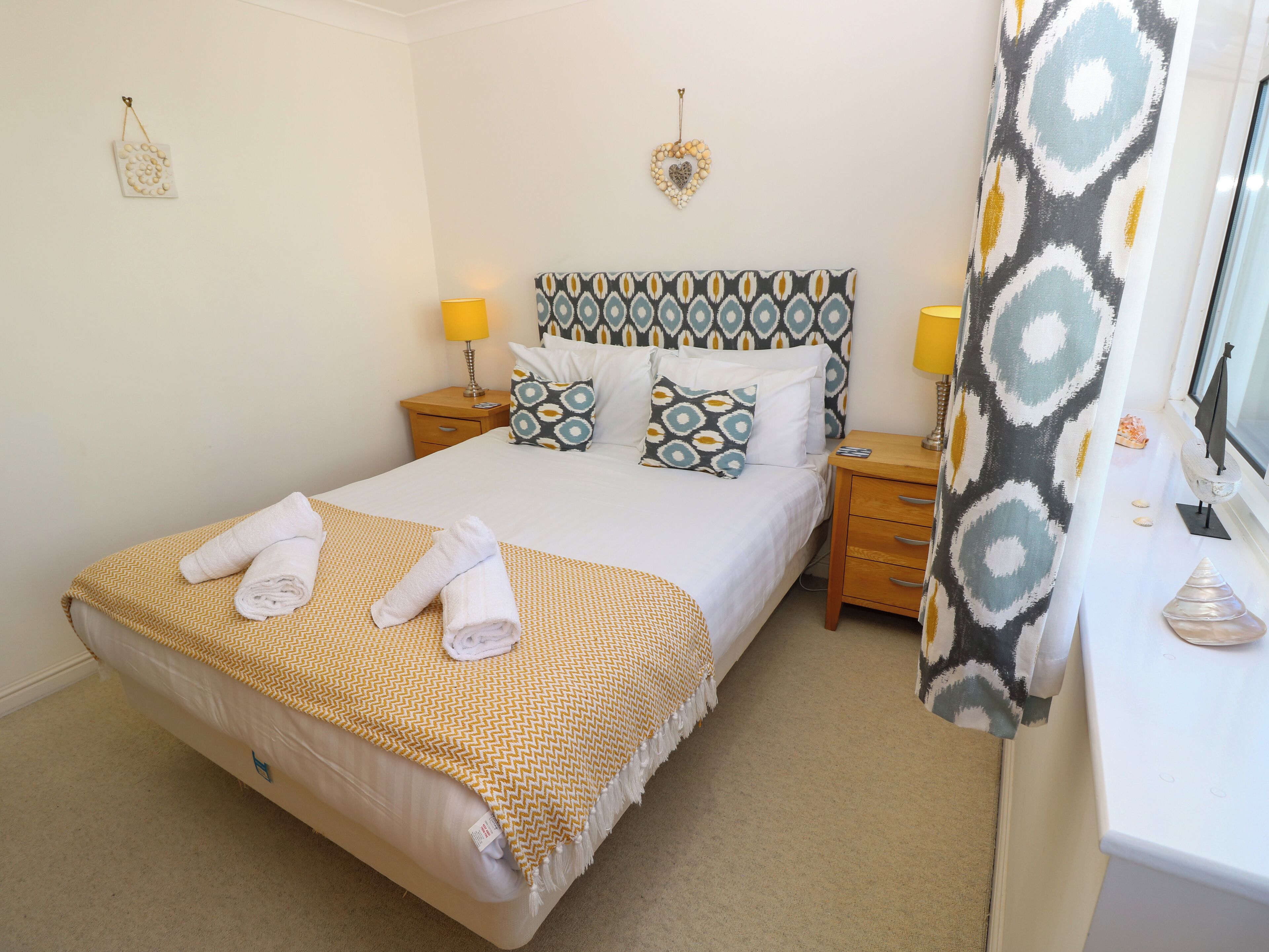 A neatly made bed with towels, a patterned headboard, two bedside lamps, and a decorative wall hanging.