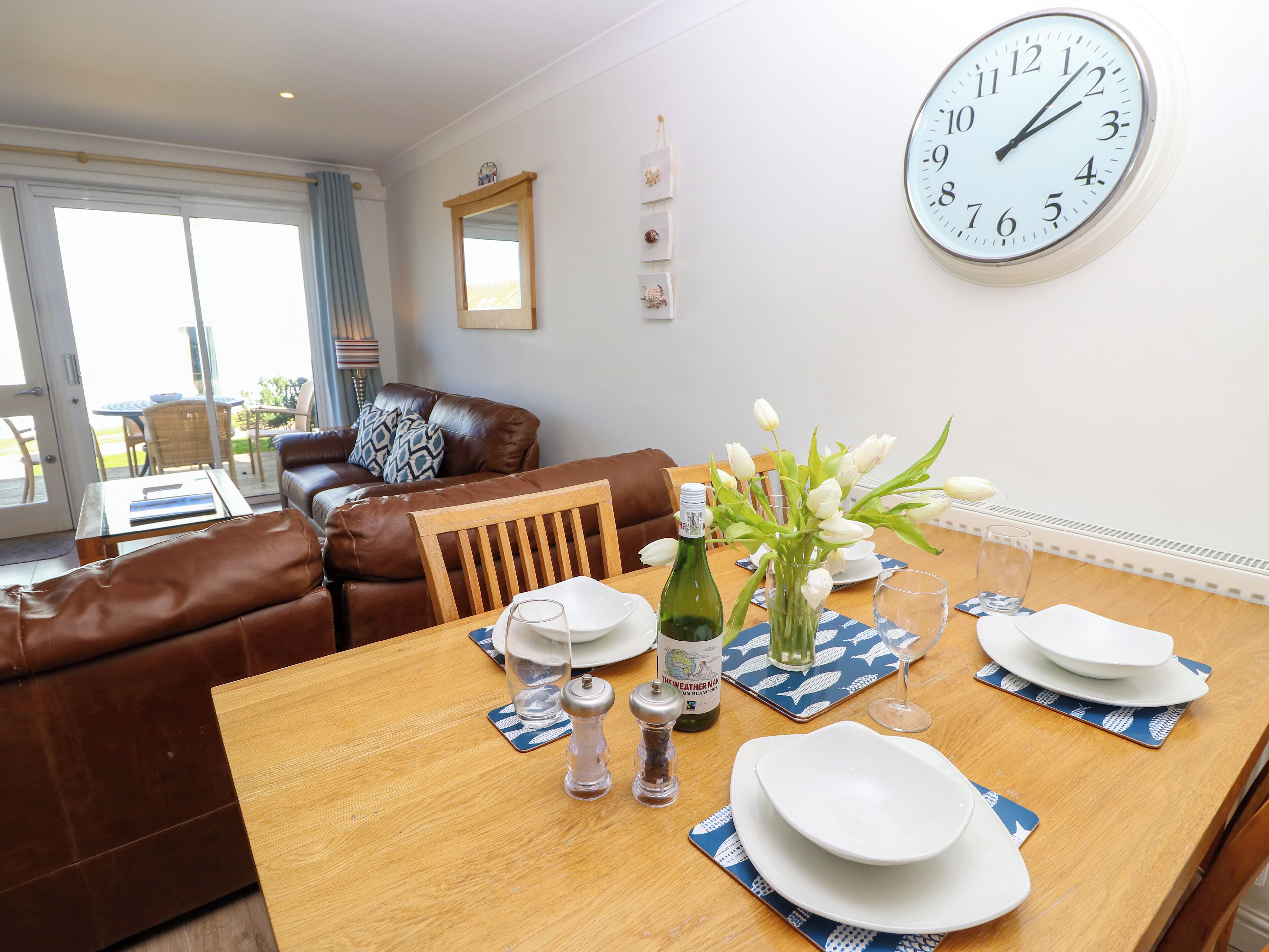A dining area with a wooden table set for two, a bottle of wine, salt and pepper shakers, and a vase with white flowers. A brown leather sofa and a large wall clock are also visible.