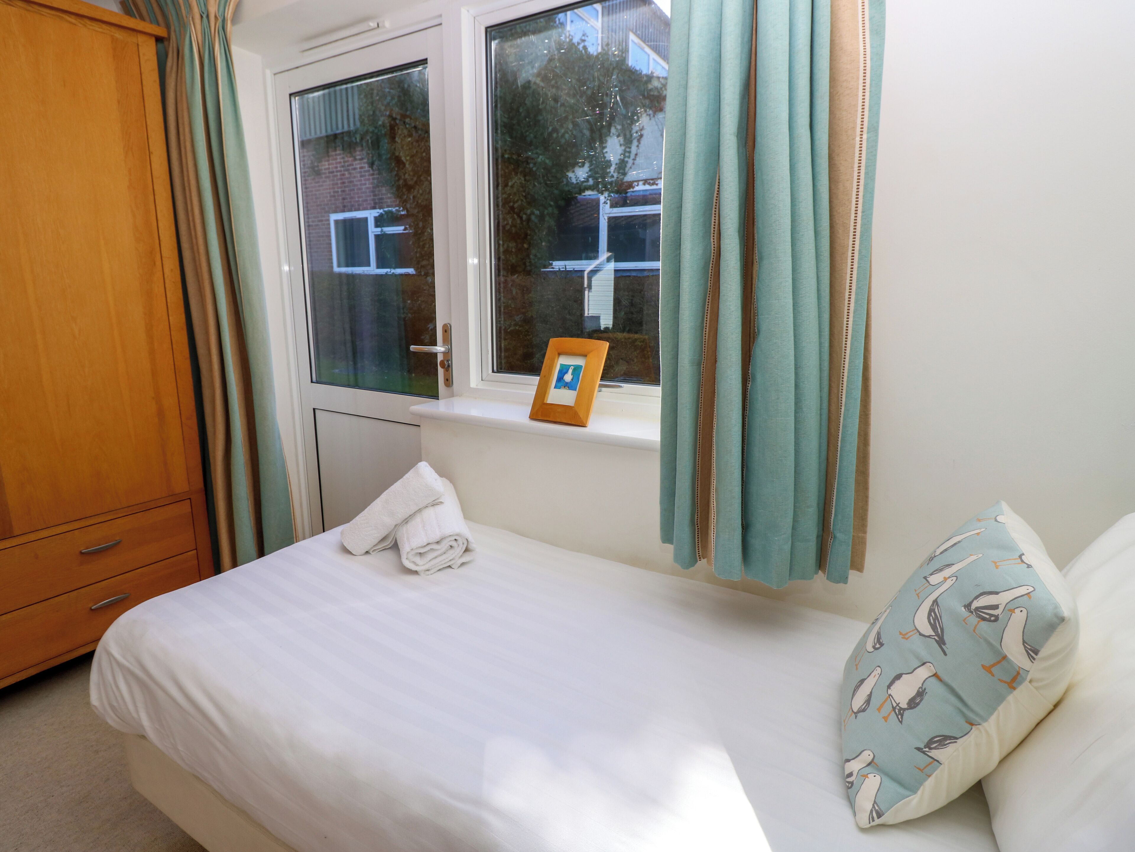 A bedroom with a bed, a window with curtains, a wooden wardrobe, and a pillow with a bird pattern.