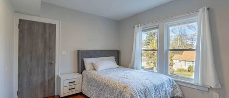 A bedroom with a bed, a nightstand, a window with curtains, and a view of the outside.