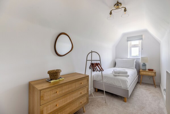 A bedroom with a wooden dresser, a bed with white linens, a hanging rack, a small table with a lamp, and a window with a striped valance.