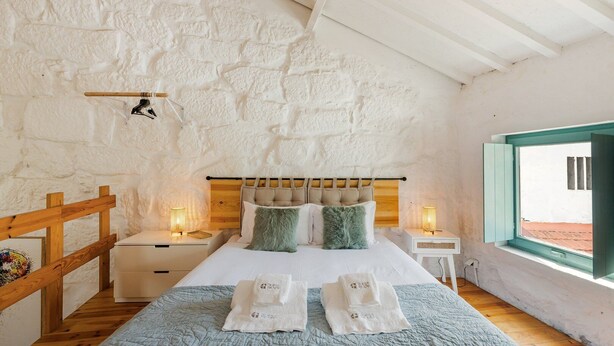 Un dormitorio con pared de piedra, cabecero de madera, dos mesitas de noche con lámparas, cama con colcha y ventana con vistas.