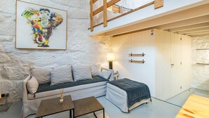 Una habitación acogedora con sofá, una mesita, una lámpara y una cama con cabecero de madera. Arriba, hay una zona tipo altillo accesible mediante una escalera de madera.