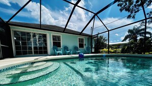 Una piscina cristalina con sillas azules y una pelota azul, rodeada por una casa con ventanas amplias y un porche cubierto.