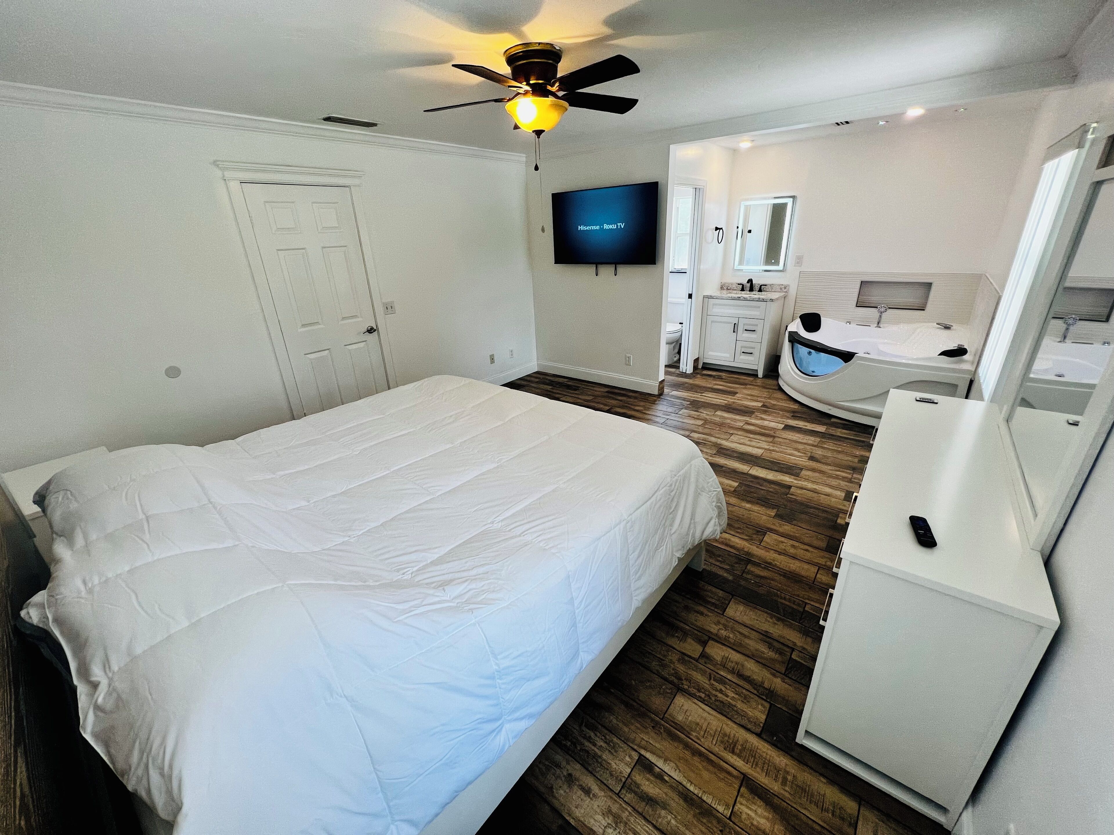 A bedroom with a bed, a ceiling fan, a TV, a bathroom with a sink and mirror, and wooden flooring.
