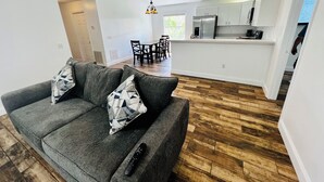 Una sala moderna con un sofá gris, cojines estampados y un control remoto en el suelo. Se ve en el fondo un comedor con sillas y una mesa.