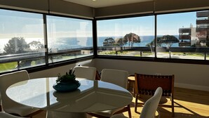 A modern room with large windows offering a view of the sea and buildings. There is a round glass table with chairs and a small plant on it. - Atrium (Cascais)
