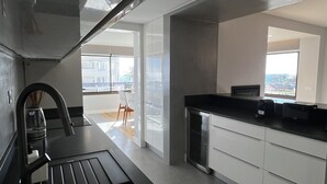 A modern kitchen with a black countertop, white cabinets, and a view of the city through large windows. - Atrium (Cascais)