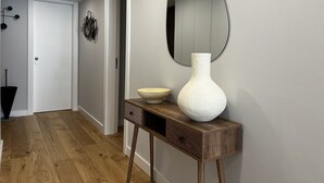 A hallway with a wooden floor, a white door, a mirror, and a wooden console table with a vase and bowl. - Atrium (Cascais)