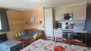 A compact kitchen with a microwave, oven, and dishwasher. There's a dining table with a tablecloth and chairs. A sofa is against the wooden wall, and a small shelf is above it. - 100m from ski slopes, 1st floor, balcony, ski locker, 48m², Valfréjus (Valfréjus)