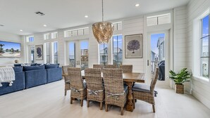 A spacious dining area with a large wooden table and wicker chairs, surrounded by large windows offering views of the neighborhood. - Elegant Beach House with views, pool, ping pong, shuffleboard - steps to beach (Port Aransas)