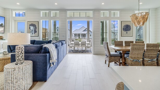 A spacious living room with a blue sofa, a dining area with wicker chairs, and a large window overlooking a house. - Elegant Beach House with views, pool, ping pong, shuffleboard - steps to beach (Port Aransas)