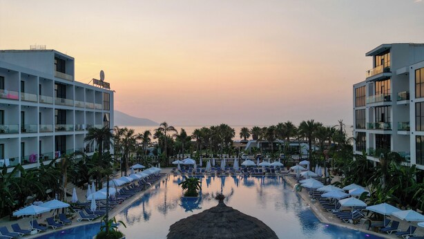 A resort pool with lounge chairs and umbrellas, surrounded by palm trees and modern buildings. - Oricon Coast (Orikum)