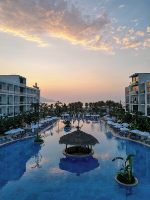 A resort pool with lounge chairs and umbrellas, surrounded by palm trees and modern buildings.