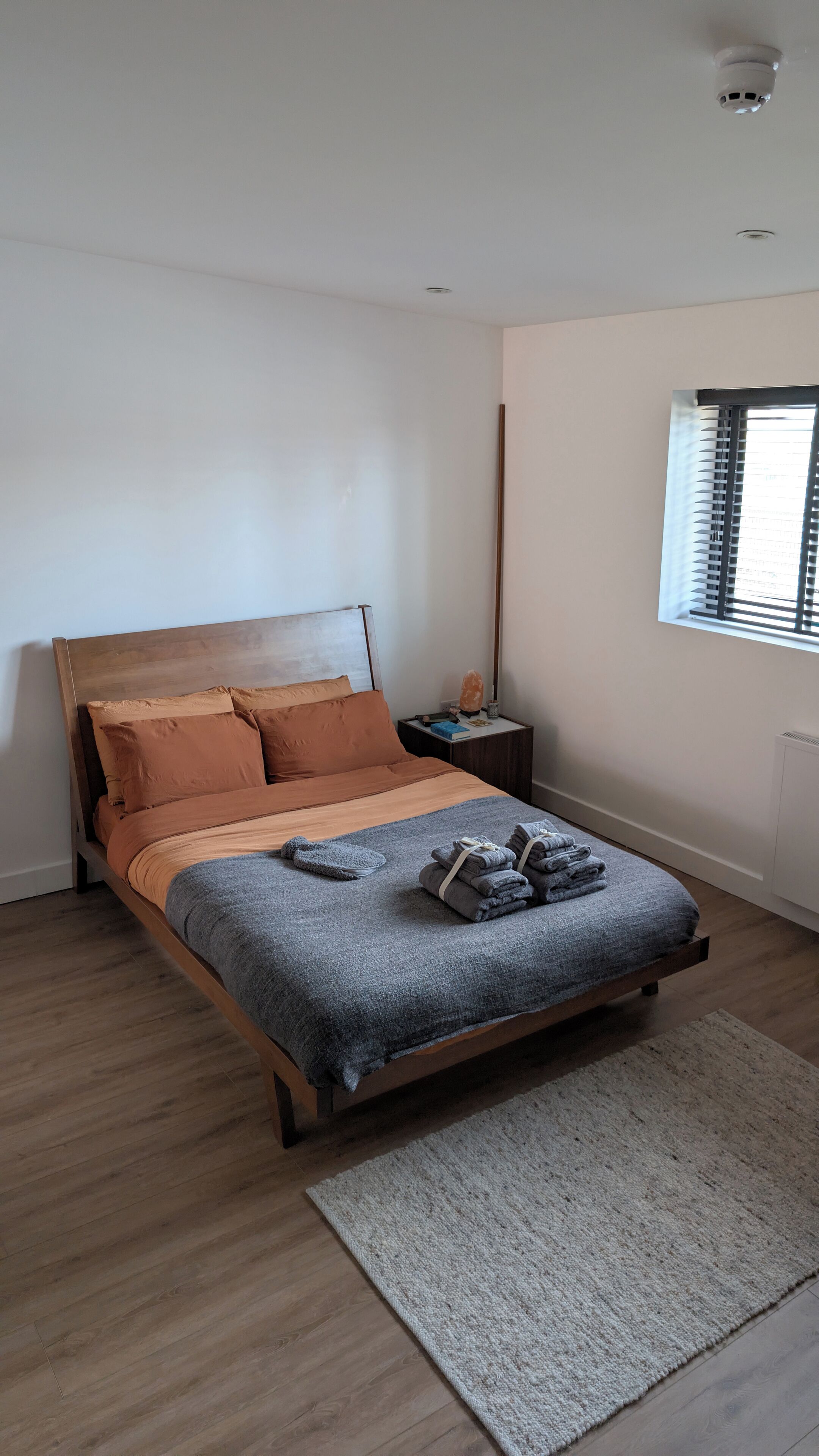 A bedroom with a wooden bed, a nightstand with a salt lamp, and a view of the outside through a window with blinds.