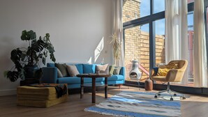 En moderne stue med en blå sofa, en gul lænestol, et sofabord og et stort vindue med udsigt til en murstensbygning.