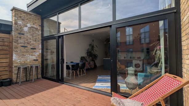 En moderne terrasse med træterrasse, et spiseområde med stole og bord, samt en skydedør ind til et rum med vaskemaskine og en stor plante.