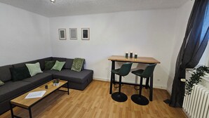 A modern living room with a dark grey sofa, a wooden coffee table, and a dining table with two chairs. There are three framed pictures on the wall, a ceiling light fixture, and a radiator on the right. - Large modern vacation apartment (Kassel)