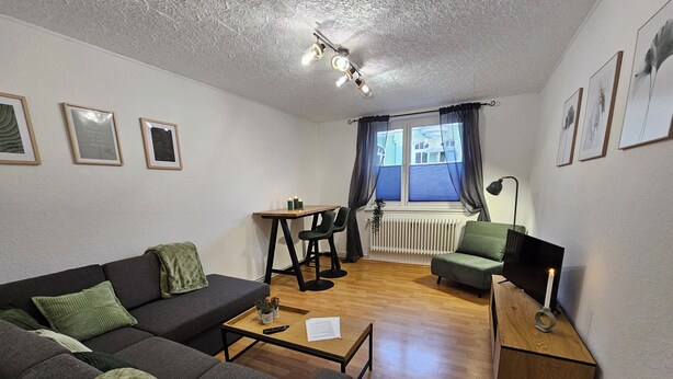 A modern living room with a sofa, a coffee table, a small table with candles, a chair, and a lamp. There are framed pictures on the wall and a window with curtains. - Large modern vacation apartment (Kassel)
