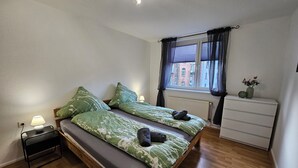 A bedroom with a wooden bed, two bedside lamps, a window with curtains, a radiator, a dresser, and a small clock. - Large modern vacation apartment (Kassel)