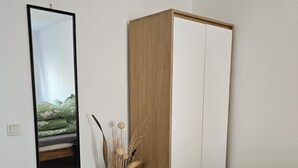 A room with a wooden wardrobe, a green chair, a white vase with dried plants, and a mirror on the wall. - Large modern vacation apartment (Kassel)