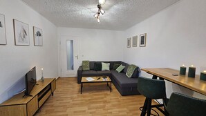 A modern living room with a wooden floor, a grey sofa with green cushions, a wooden coffee table, a TV on a wooden stand, and a ceiling light fixture. - Large modern vacation apartment (Kassel)