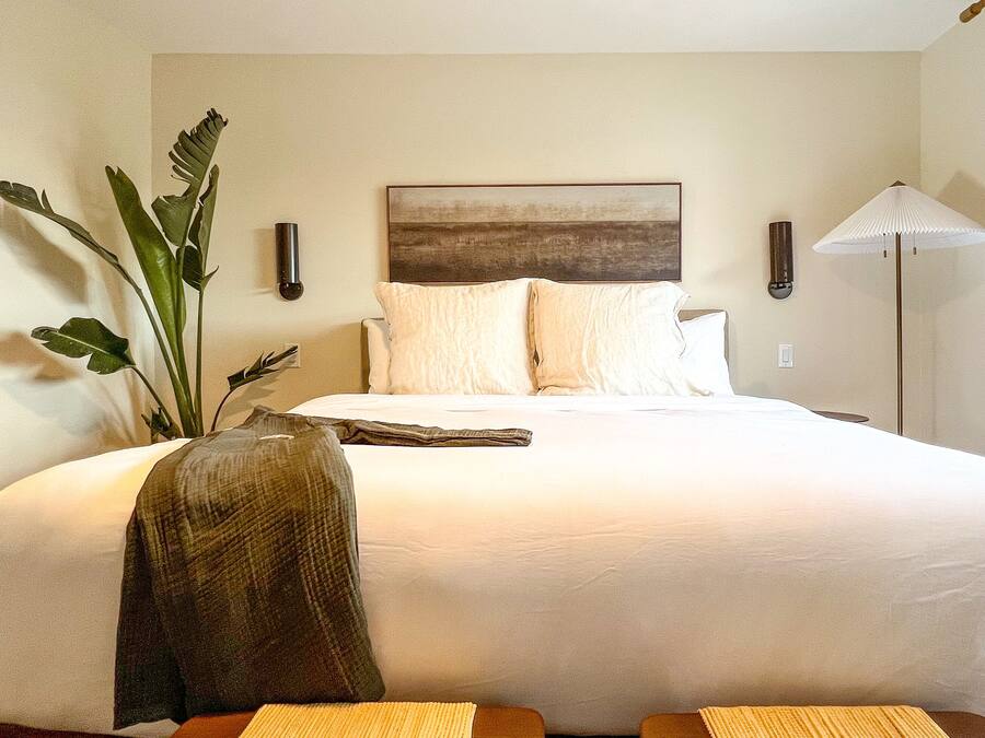 A hotel room with a large bed, a painting on the wall, a floor lamp, and a plant.