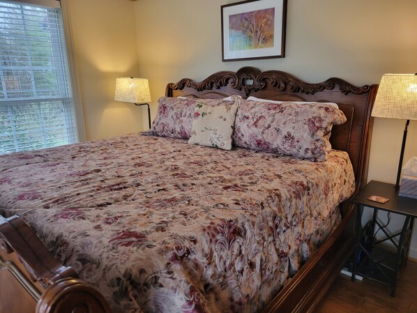 A bed with a floral bedspread, two bedside lamps, a framed picture on the wall, and a window with blinds. - Charming 3-bedroom home in Laurel Springs with view over the Blue Ridge Parkway (Laurel Springs)