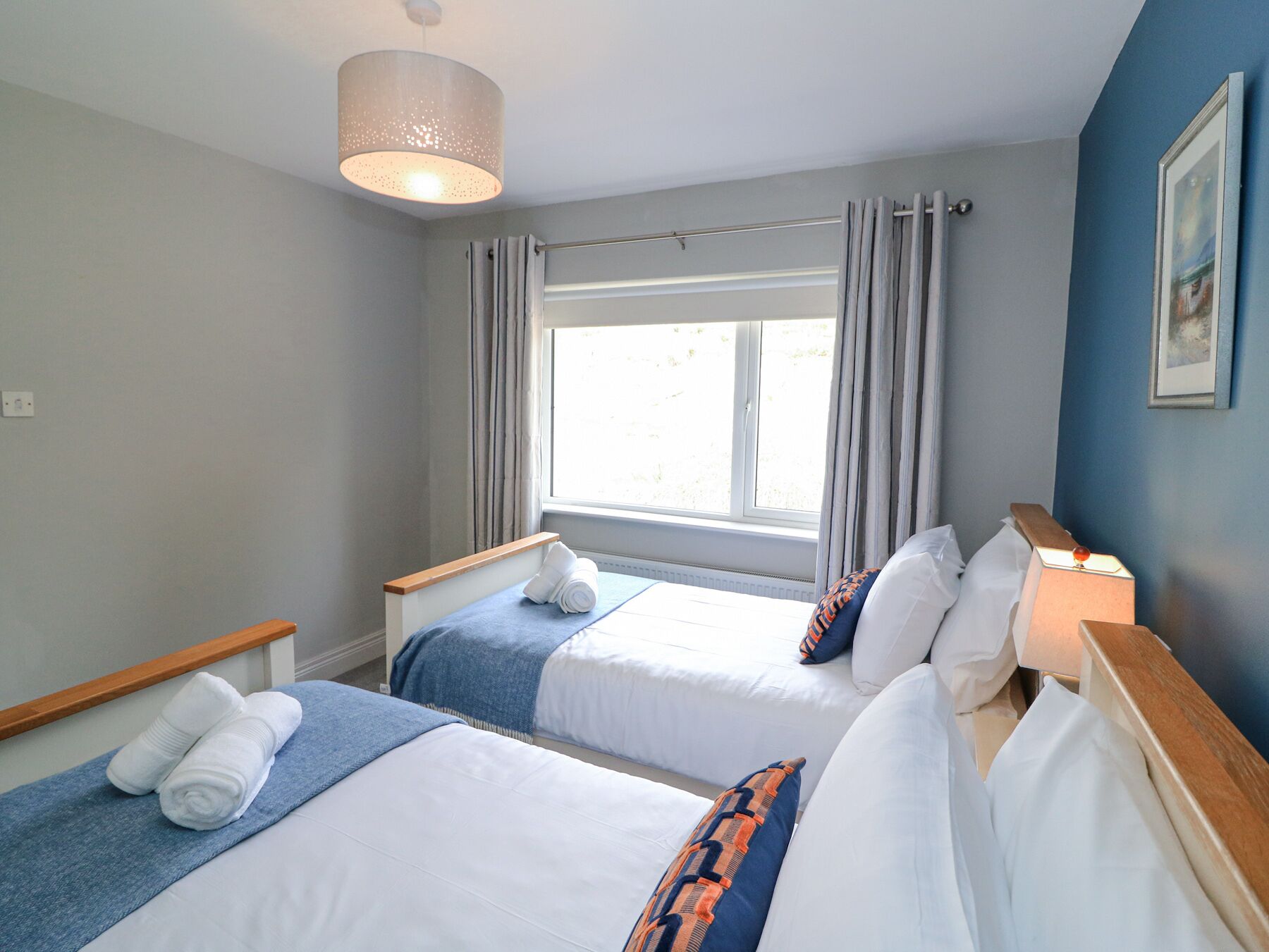 Habitación de hotel con dos camas, una ventana con cortinas, una lámpara colgante y un cuadro en la pared.