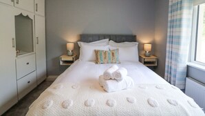 A neatly made bed with white linens and rolled towels, two bedside lamps, a headboard, and a window with curtains.