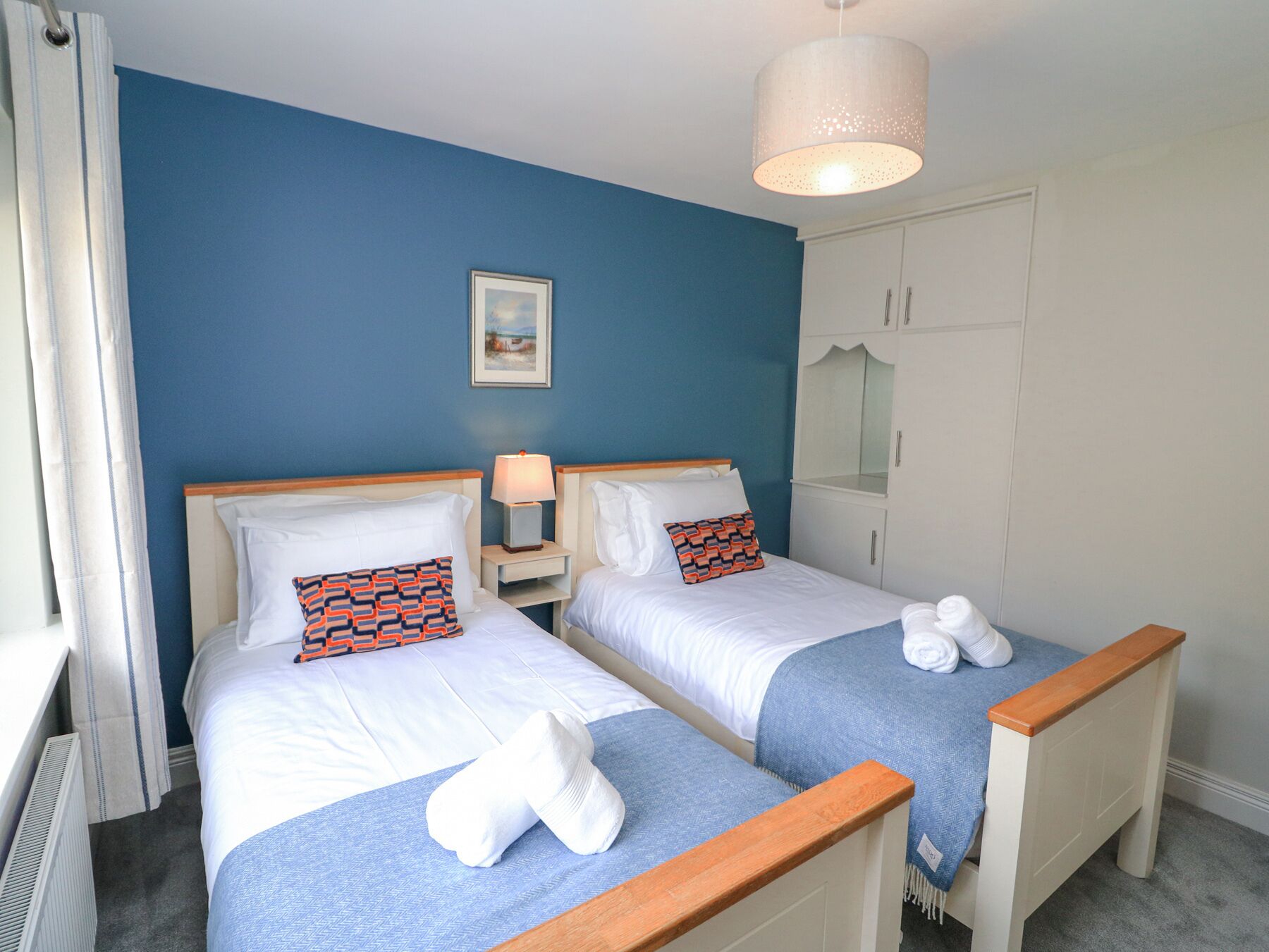 Habitación de hotel con dos camas, un armario blanco, una mesita de noche con lámpara y un cuadro en la pared.