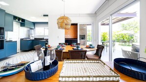 A modern kitchen with blue cabinets, a dining area with a large table, wicker chairs, and a hanging light fixture. - Bz83 Casa no Centro Piscina e 4 suítes (Buzios)