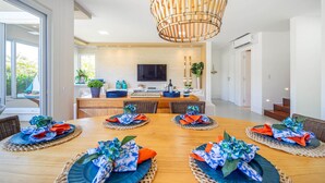A modern dining area with a wooden table set for six, a large woven light fixture above, and a kitchen in the background. - Bz83 Casa no Centro Piscina e 4 suítes (Buzios)