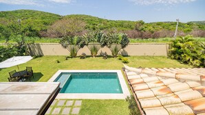 A swimming pool with a tiled deck, surrounded by a well-maintained lawn and various plants. - Bz83 Casa no Centro Piscina e 4 suítes (Buzios)