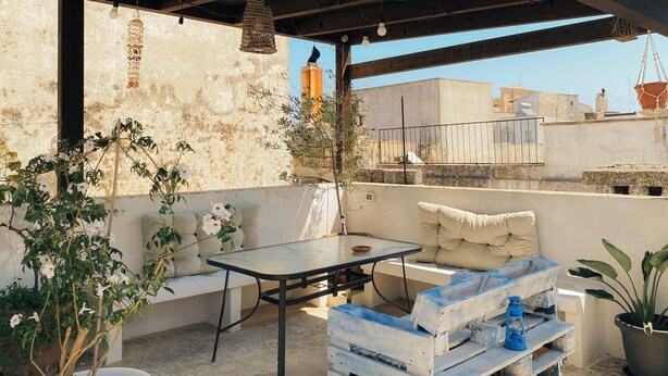 Pátio externo com mesa e bancos, cercado por vasos de plantas e um lustre pendurado.