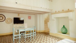 Um quarto com televisão, uma mesa e cadeiras, um chão estampado, uma escada e um vaso verde numa prateleira.