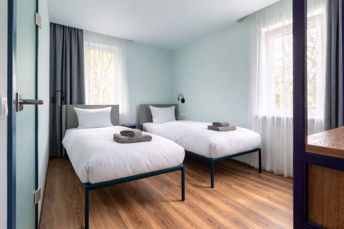 A hotel room with two beds, a wooden floor, and a view of trees through the window.