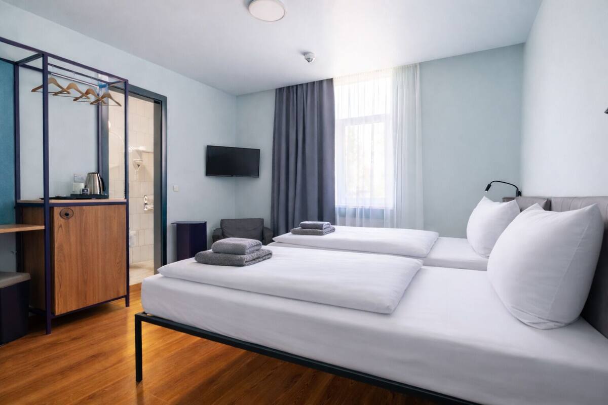 A modern hotel room with a bed, a TV, a wardrobe, and a window with curtains.