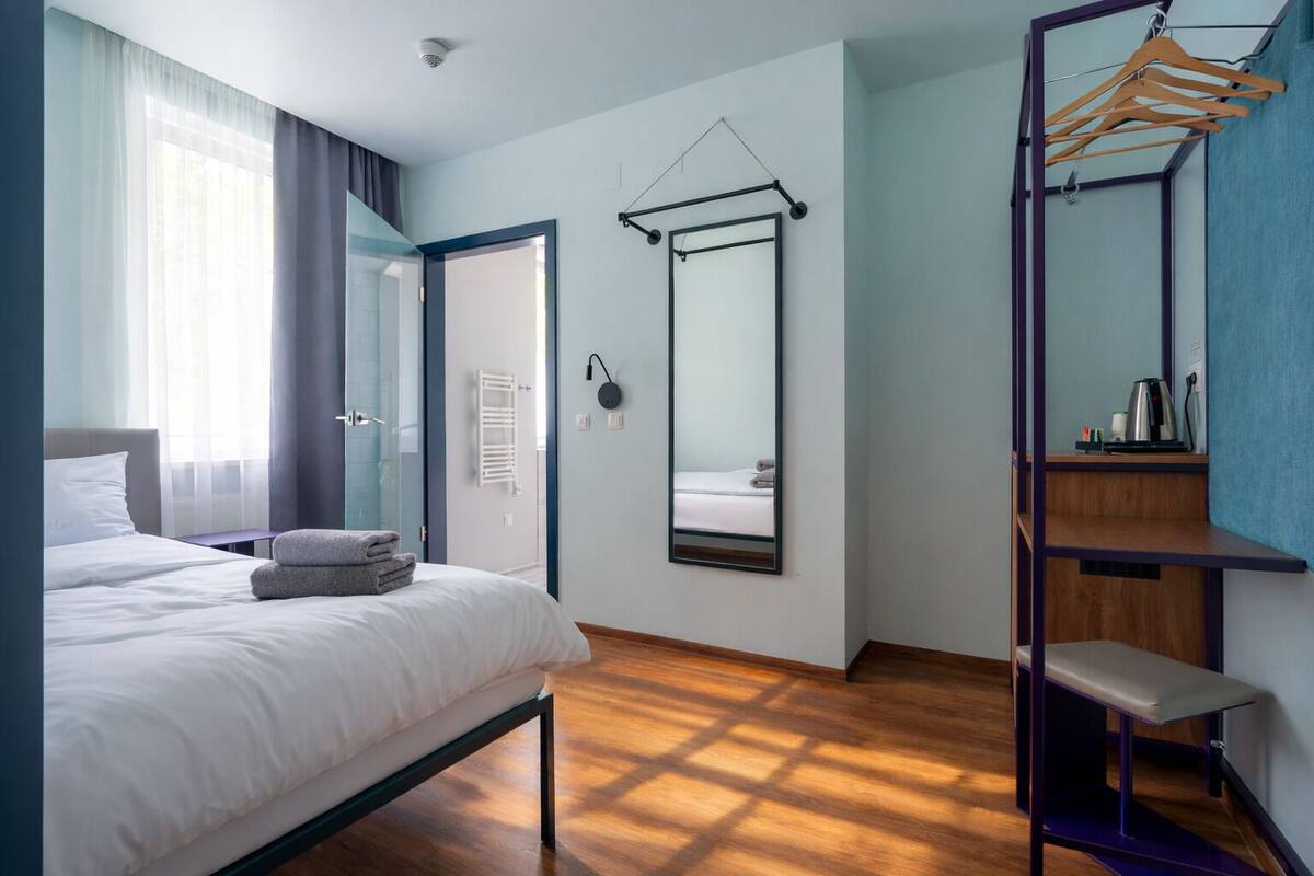 A modern hotel room with a bed, a mirror, a wooden shelf, and a wardrobe.