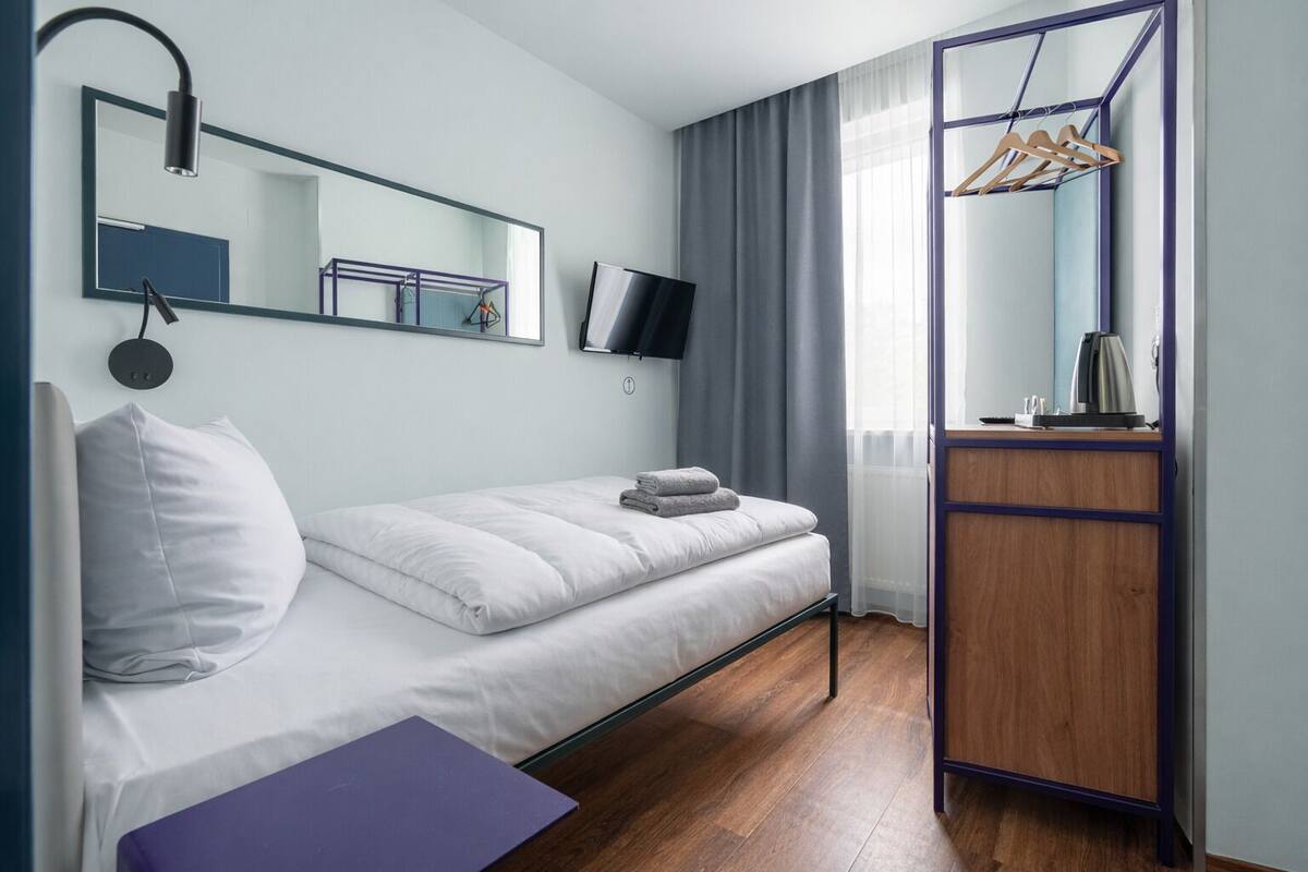 A modern hotel room with a single bed, a wooden wardrobe, a desk, and a TV mounted on the wall.